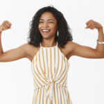 She can deal with any trouble. Portrait of strong and independet joyful stylish african american sportswoman working out in gym standing in trendy overalls and showing muscles winking and smiling