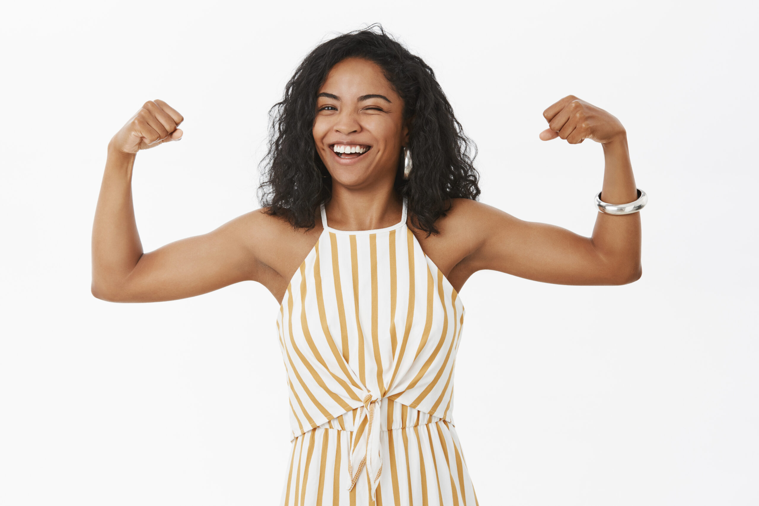 She can deal with any trouble. Portrait of strong and independet joyful stylish african american sportswoman working out in gym standing in trendy overalls and showing muscles winking and smiling